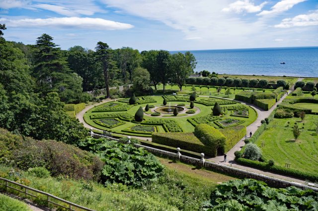 A scenic view of a formal garden with geometric hedges, a central fountain, and a backdrop of the sea under a blue sky.