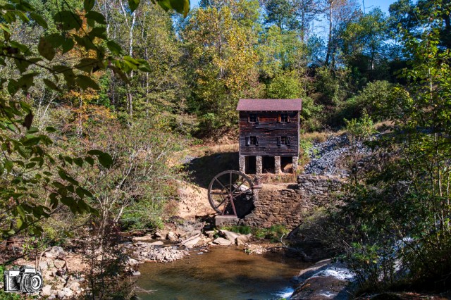 Historic watermill surrounded by greenery, featuring a large water wheel, with a stream flowing in front.