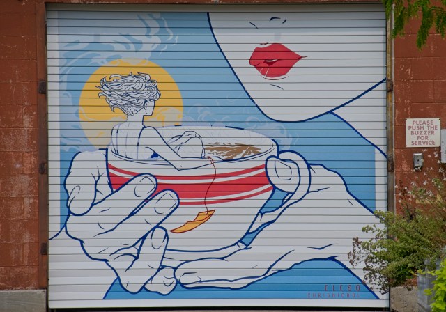 A colorful mural depicting a person sitting in a large bowl while holding it, with a sun in the background and a close-up of a red-lipped mouth.
