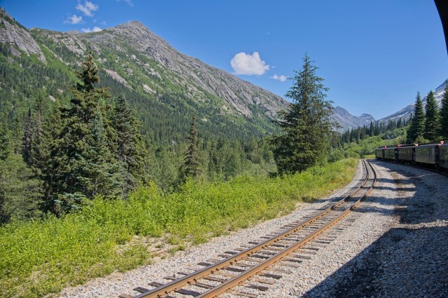 Scenic view of train tracks winding through a lush green valley surrounded by mountains and trees under a bright blue sky.