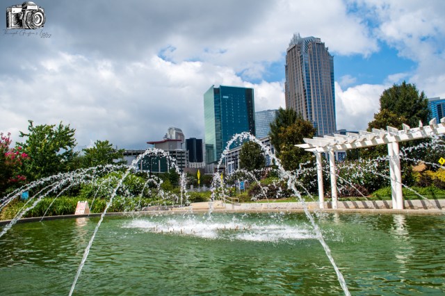 A city park fountain with water jets, surrounded by greenery and modern skyscrapers against a partly cloudy sky.