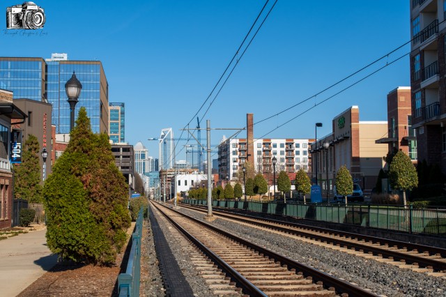 View of railway tracks lined with greenery and buildings in an urban setting, under a clear blue sky.