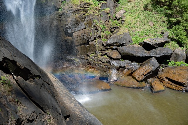 A waterfall cascading over rocky cliffs into a serene pool below, with a faint rainbow appearing in the mist.