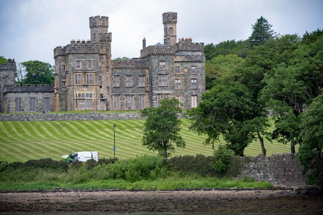 A historic castle with turrets and ornate architecture, surrounded by neatly trimmed lawns and trees. A white van is parked on the grounds next to a lamp post.