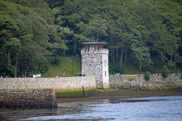 A stone watchtower beside a waterfront, surrounded by lush greenery and a walking path with people.