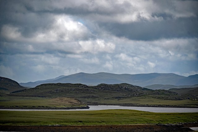 A panoramic view of rolling green hills and a calm body of water under a cloudy sky, with distant mountains visible on the horizon.
