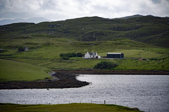 A scenic view of a white house and a nearby barn situated on green hills next to a calm body of water, with mountains visible in the background.