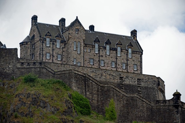 Historic stone castle situated on a rocky hill, featuring multiple gabled rooftops and large windows against a cloudy sky.