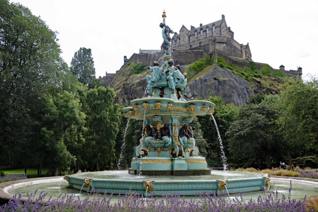 A decorative fountain in a lush park with a backdrop of a historic castle perched on a rocky hill.
