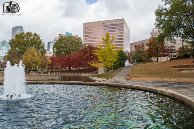 A city park featuring a tranquil pond with a fountain, surrounded by trees showcasing autumn colors, with modern buildings in the background under a cloudy sky.