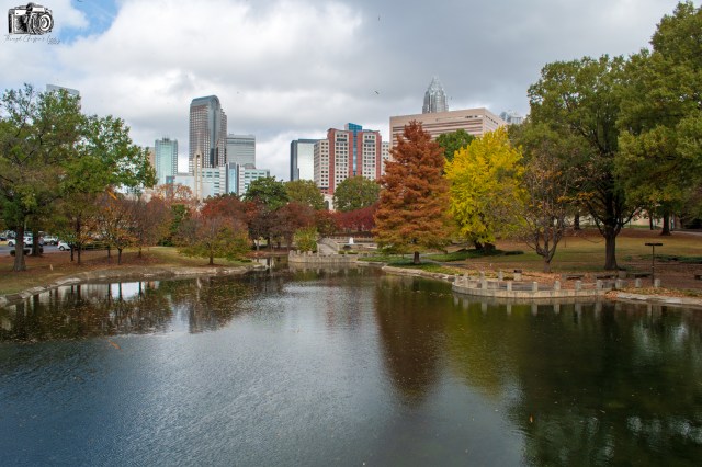A serene park view featuring a small pond surrounded by trees with autumn foliage, and a skyline of tall buildings in the background under a partly cloudy sky.