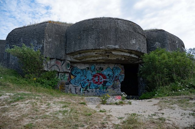 An abandoned concrete bunker covered in graffiti, surrounded by greenery and grass under a cloudy sky.