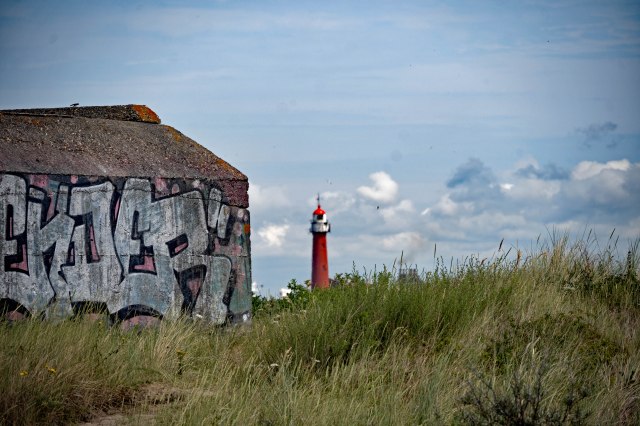 A graffiti-covered concrete structure in the foreground with a red and white lighthouse visible in the background against a cloudy sky.