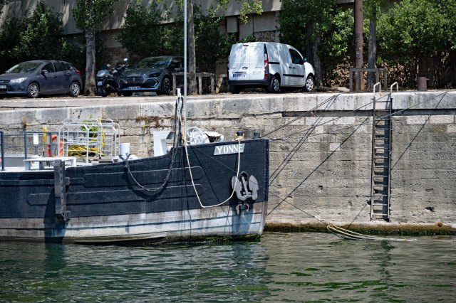 A black boat named 'Yonne' docked at a waterfront with vehicles parked nearby and a metal ladder leading up to the shore.