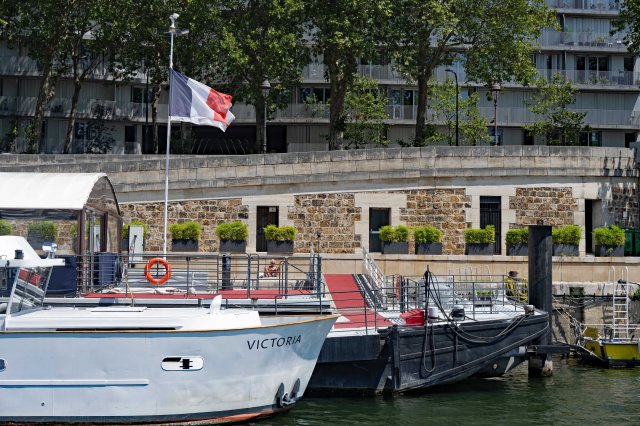 A scenic view of boats docked along a waterfront, featuring a white boat named 'VICTORIA' and a flag of France waving in the breeze. The background includes greenery and a modern building facade.